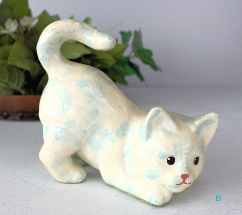 K&ouml;nnte beinhalten: Cremefarbene Katzenfigur mit hellblauem Blumenmuster. Die Katze befindet sich in einer spielerischen Pose, mit gebogenem Schwanz und gew&ouml;lbtem K&ouml;rper. Die Katze hat eine rote Nase und schwarze Augen.