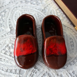 Pode incluir: Dois tamancos de cerâmica, cada um com um acabamento brilhante. A metade superior de cada tamanco é vermelha vibrante, passando para um marrom escuro na base. Estes tamancos decorativos são provavelmente para exibição ou pequenas plantas.