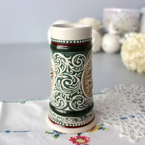 Op de afbeelding: Een groene en witte keramische bierkan met een gedetailleerd bloemenpatroon en een bruin hertenhoofd. De kan heeft een rode band rond de bovenkant en de woorden "Art Pottery" aan de onderkant.