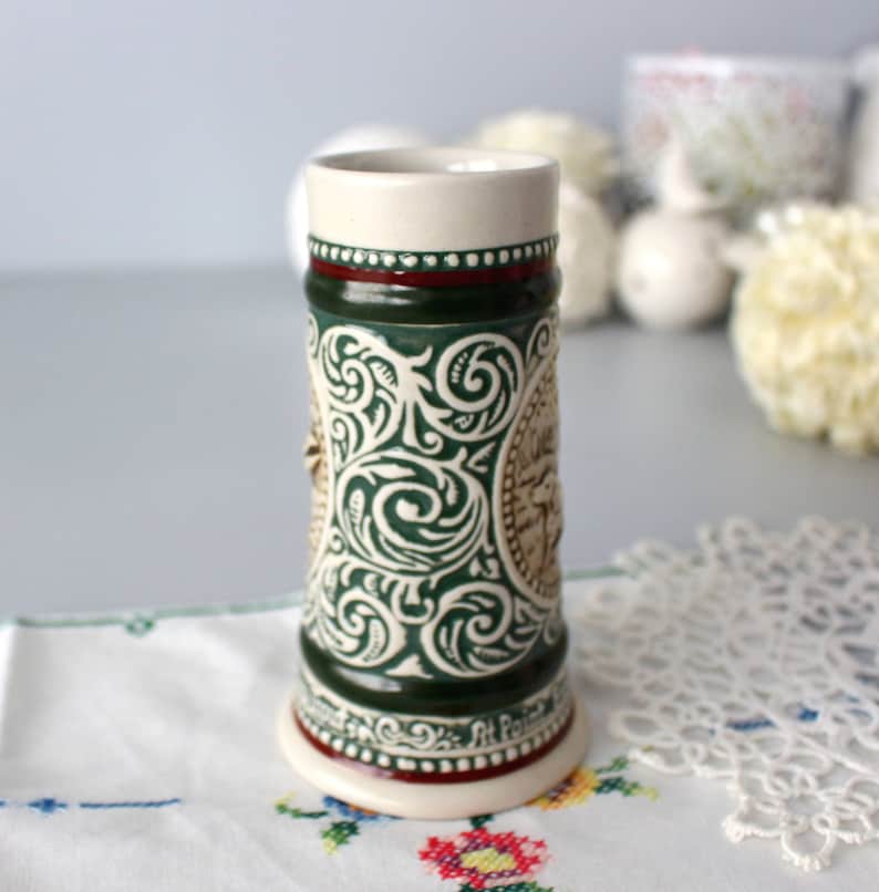 Op de afbeelding: Een groene en witte keramische bierkan met een gedetailleerd bloemenpatroon en een bruin hertenhoofd. De kan heeft een rode band rond de bovenkant en de woorden "Art Pottery" aan de onderkant.