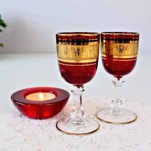 Könnte beinhalten: Zwei rote Glas-Schnapsgläser mit goldenen Verzierungen und klaren Stielen. Die Gläser stehen auf einer weißen Spitzenunterlage mit einem roten Glas-Kerzenhalter im Vordergrund.