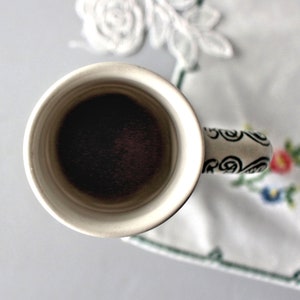 Op de afbeelding: Een witte keramische mok met een zwart-wit swirl-ontwerp op de handgreep. De mok is gevuld met donkere koffie.