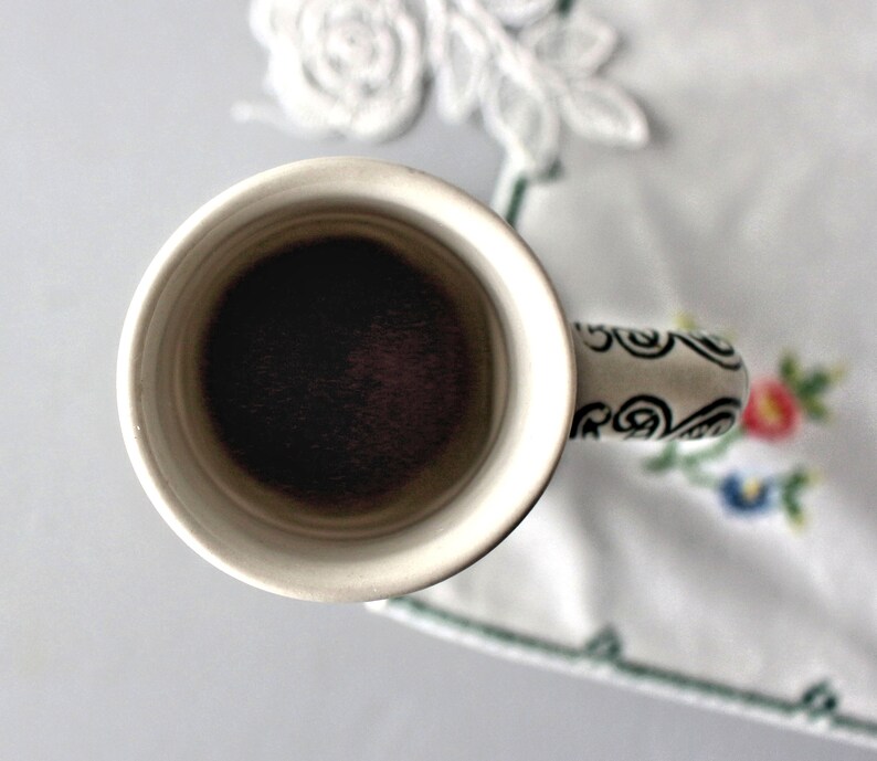 Op de afbeelding: Een witte keramische mok met een zwart-wit swirl-ontwerp op de handgreep. De mok is gevuld met donkere koffie.
