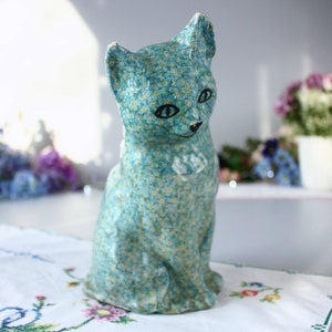 Pode incluir: Uma figura de cer&acirc;mica azul de um gato com um padr&atilde;o de flores de miosotis. O gato est&aacute; sentado com a cabe&ccedil;a ligeiramente inclinada para o lado.