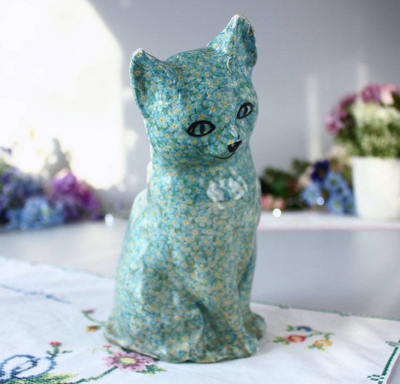 Pode incluir: Uma figura de cer&acirc;mica azul de um gato com um padr&atilde;o de flores de miosotis. O gato est&aacute; sentado com a cabe&ccedil;a ligeiramente inclinada para o lado.