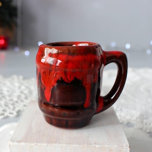 May include: A small, brown ceramic mug with a handle. The top half of the mug is covered in a vibrant, dripping red glaze. The mug is sitting on a white surface.