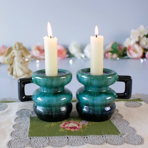 May include: Two teal ceramic candle holders with black handles, each holding a lit, ivory-coloured candle. The holders sit on a green embroidered placemat with a floral design, with a decorative angel figurine in the background.
