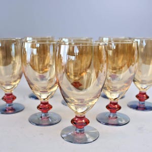 May include: A collection of eight vintage wine glasses. Each glass features a golden-amber bowl, a red stem, and a blue base. The glasses are displayed on a white surface, reflecting light and creating a warm aesthetic.