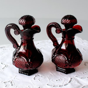 Puede incluir: Dos aceiteras de vidrio rojo rubí con asas ornamentadas y tapones texturizados. Las aceiteras tienen un cuerpo bulboso con un patrón decorativo y una base cuadrada. El fondo es un mantel blanco y de encaje.