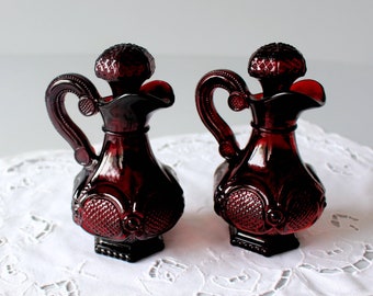 May include: Two ruby red glass cruets with ornate handles and textured stoppers. The cruets have a bulbous body with a decorative pattern and a square base. The background is a white, lacy tablecloth.