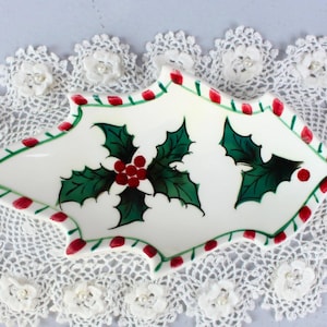Könnte beinhalten: Eine weiße Keramikschale mit Stechpalmenbeeren-Design, mit grünen Blättern und roten Beeren, liegt auf einer weißen Spitzendecke. Die Schale hat einen rot-grünen Rand. Dekorativer Artikel zum Thema Weihnachten.
