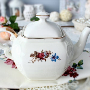 May include: White porcelain teapot with a gold trim and floral design. The teapot has a square body and a lid with a knob. The floral design features red roses, blue flowers, and green leaves.