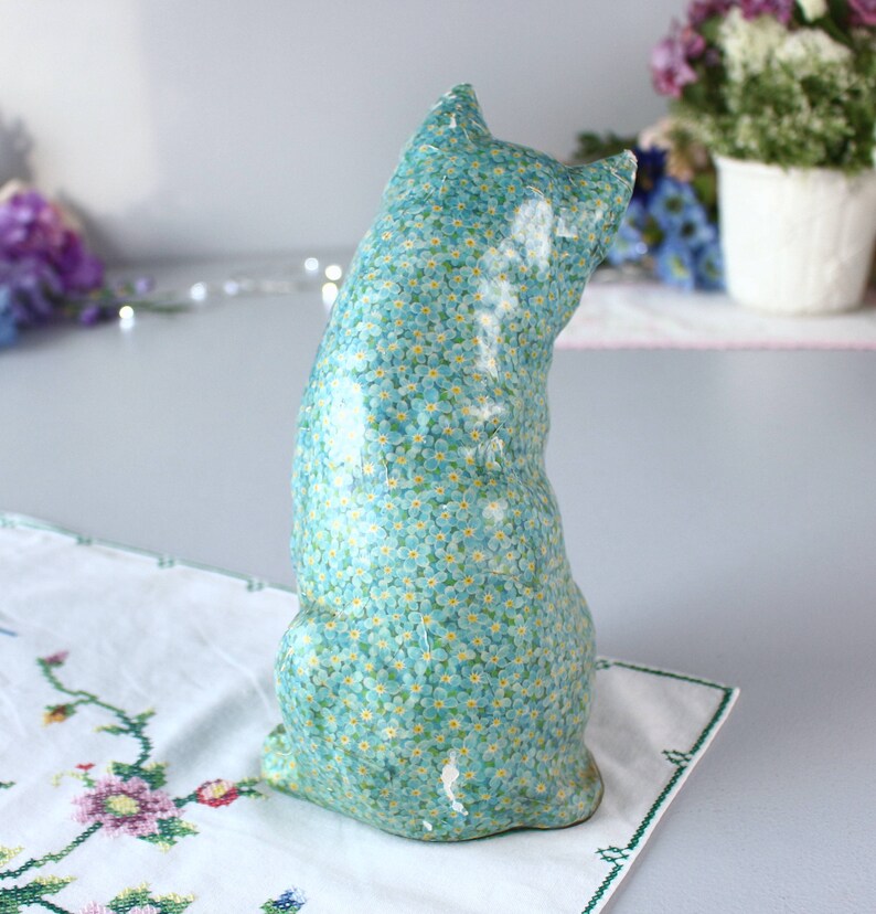 Pode incluir: Uma figura de cer&acirc;mica azul de um gato com um padr&atilde;o de flores de miosotis. O gato est&aacute; sentado com o rabo enrolado em torno do seu corpo.