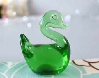 May include: A small, emerald green glass swan figurine. The swan has a curved neck and a detailed beak. The figurine is sitting on a light blue and white patterned surface. The glass is transparent, allowing light to pass through.