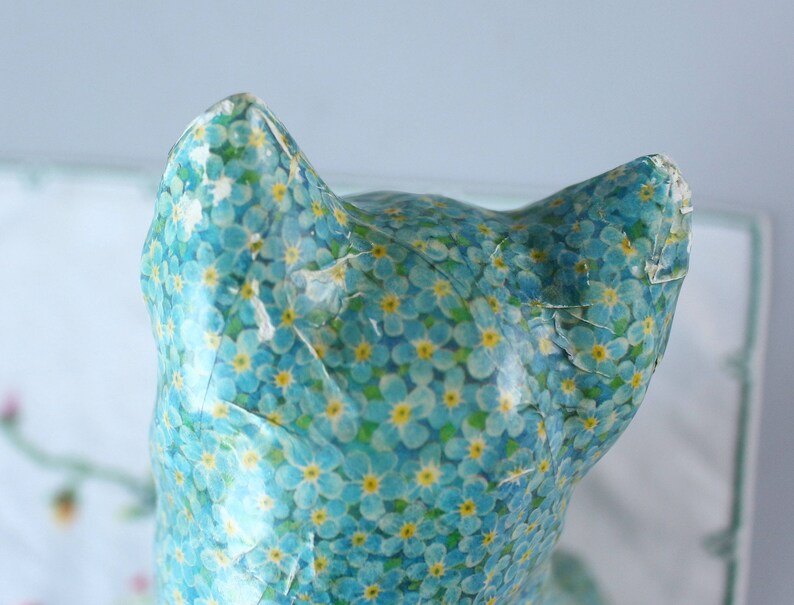 Pode incluir: Uma figura de cer&acirc;mica de um gato coberta com um padr&atilde;o floral azul e amarelo. O gato est&aacute; de costas para a c&acirc;mera e tem uma cauda longa.