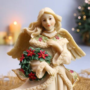Puede incluir: Una figura de ángel color crema con cabello rubio y alas emplumadas. El ángel viste una túnica color crema con un cuello decorativo y sostiene una corona verde adornada con poinsettias rojas. La figura tiene una expresión serena y es una decoración navideña.