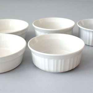 May include: Six white ceramic ramekins of varying sizes are arranged on a light grey surface. Some have a smooth exterior, while others feature vertical ribbed detailing. These small, oven-safe dishes are ideal for baking individual portions.
