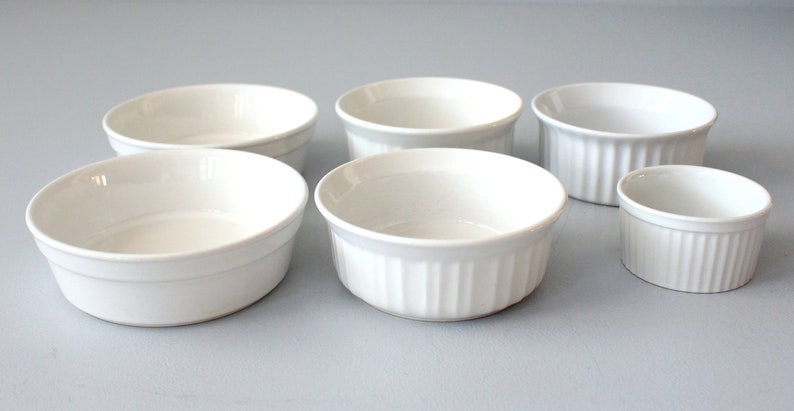 May include: Six white ceramic ramekins of varying sizes are arranged on a light grey surface. Some have a smooth exterior, while others feature vertical ribbed detailing. These small, oven-safe dishes are ideal for baking individual portions.