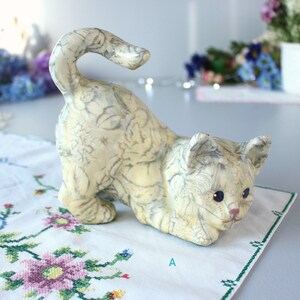 K&ouml;nnte beinhalten: Eine cremefarbene Keramik-Katzenfigur in spielerischer Pose mit einem grauen Blumenmuster. Die Katze hat blaue Augen und eine rosa Nase und liegt auf einem wei&szlig;en, bestickten Tuch.