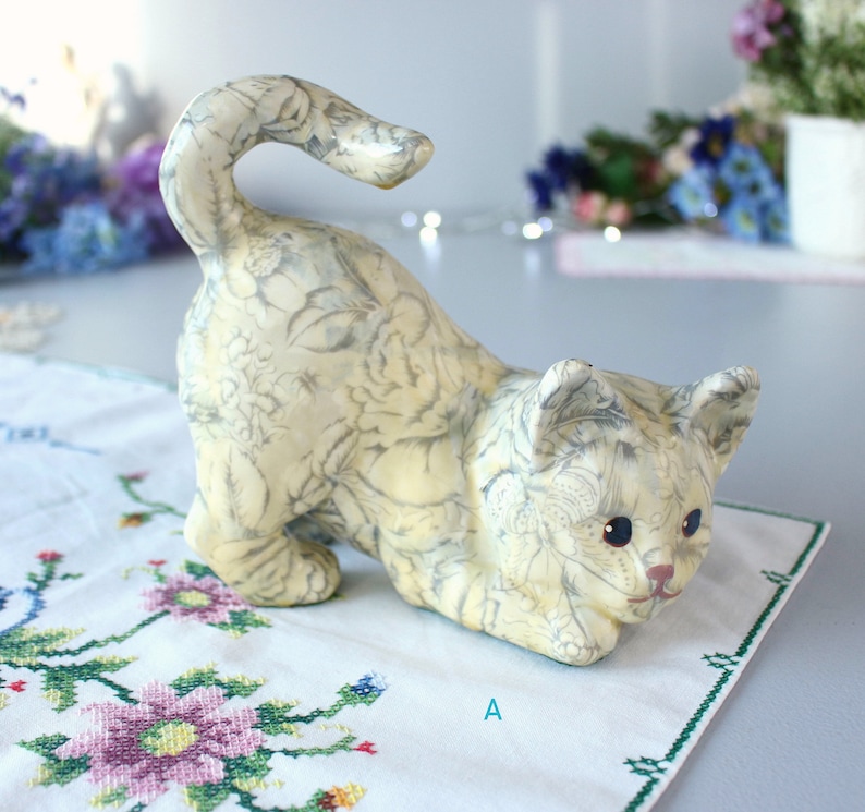 K&ouml;nnte beinhalten: Eine cremefarbene Keramik-Katzenfigur in spielerischer Pose mit einem grauen Blumenmuster. Die Katze hat blaue Augen und eine rosa Nase und liegt auf einem wei&szlig;en, bestickten Tuch.