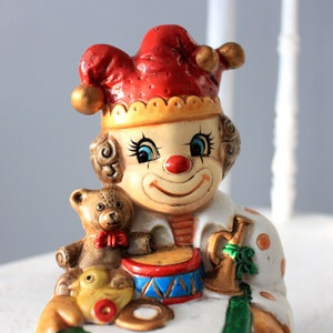 May include: A ceramic clown figurine wearing a red and yellow hat, with blue eyes and a red nose. The clown is holding a toy drum and a teddy bear.