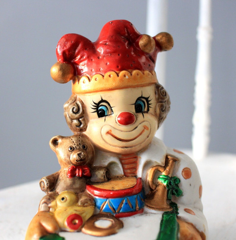 May include: A ceramic clown figurine wearing a red and yellow hat, with blue eyes and a red nose. The clown is holding a toy drum and a teddy bear.