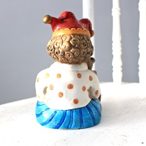 May include: A ceramic figurine of a jester with a red and yellow hat, brown curly hair, a white shirt with orange polka dots, and a blue skirt with white stripes.
