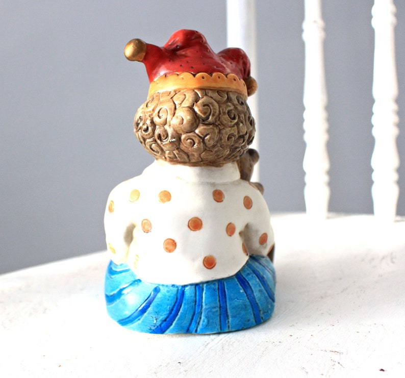 May include: A ceramic figurine of a jester with a red and yellow hat, brown curly hair, a white shirt with orange polka dots, and a blue skirt with white stripes.