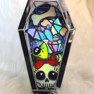 May include: A stained glass coffin-shaped display piece featuring a central skull with a red bow, a green spider, a purple bat, a bone, and a diamond shape. The frame is made of dark metal, and the interior contains iridescent and colorful glass.