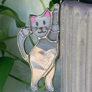 May include: A stained glass cat ornament featuring white and grey marbled glass pieces held together by silver metal solder lines. The cat has small pink painted ears, a brown painted face, and a curved tail, designed to hang from a vertical edge.