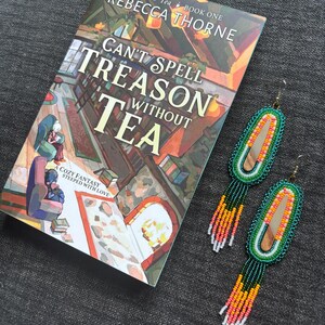 May include: A paperback book titled Can't Spell Treason Without Tea by Rebecca Thorne, featuring an illustrated interior scene, alongside a pair of long, oval-shaped beaded earrings with green, orange, pink, and white fringe detailing.