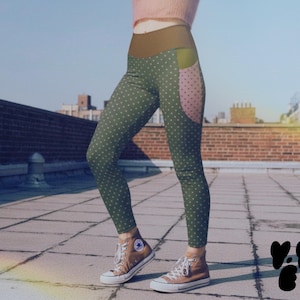 Puede incluir: Leggings verdes con lunares blancos y un bolsillo rosa. Los leggings tienen una cintura alta marrón. La modelo lleva zapatillas altas marrones. El fondo es un cielo azul y un edificio de ladrillo.