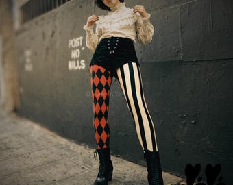 May include: A cream-colored long-sleeve blouse with a ruffled high collar, black velvet high-waisted shorts with lace-up front detail, and mismatched leggings featuring a red and black diamond pattern on one leg and black and cream stripes on the other.