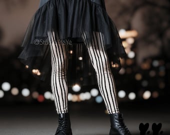 Puede incluir: Una falda negra con una capa de tul transparente con dobladillo asimétrico, combinada con leggings de color crema con rayas verticales negras y botas de cuero negro con cordones. El texto Studio YéYé es visible en la tela de la falda.