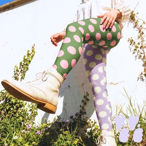 Pode incluir: Leggings com fundo verde e grandes bolinhas cor-de-rosa. A outra perna tem um fundo roxo com bolinhas brancas grandes. A pessoa está usando botas creme. O fundo é uma parede branca com plantas verdes.