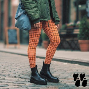 May include: Orange leggings featuring a black grid pattern, paired with a dark olive green quilted jacket, a blue denim shoulder bag, and black ankle boots with thick soles. The outfit is worn on a cobblestone street with a blurred background.