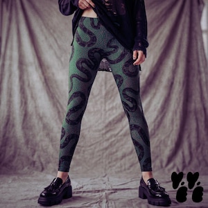 Puede incluir: Leggings con base de color verde, un patrón repetitivo de serpientes negras y pequeños puntos blancos. Los leggings se combinan con una camiseta negra de manga larga y mocasines negros de plataforma gruesa con acabado brillante.