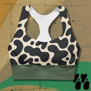 May include: A sports bra with a cow print design. The bra features a black and cream-colored pattern on the front and back, with a white racerback and a solid olive green band at the bottom. The bra is designed for athletic activities.
