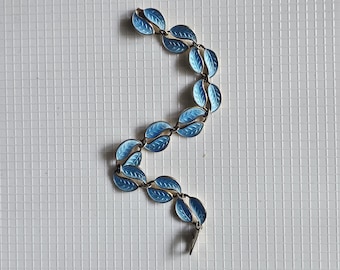 May include: A vintage bracelet featuring a series of blue enamel leaf-shaped links connected by gold-toned metal. The leaves have detailed veining, and the bracelet is laid out on a white tiled surface.