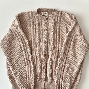 Puede incluir: Un cárdigan de punto beige con un patrón de trenza y detalles de pompón. El cárdigan tiene cierre de botón y mangas largas.