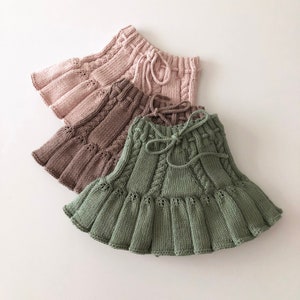 May include: Three knitted skirts in different colors: pink, brown, and green. Each skirt has a ruffled hem and a drawstring waist.