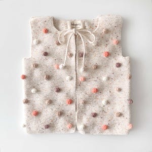 May include: Cream-colored knitted vest adorned with colorful pom-poms in shades of peach, white, and brown. The vest features a tie closure and a small label that reads "babytoly". This handmade garment is perfect for layering and adds a playful touch to any outfit.