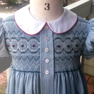 May include: A light blue smocked dress with a white collar and red trim. The dress has a button-up front and puffed sleeves. The dress is embroidered with red flowers and black stitching.