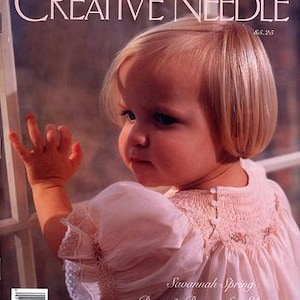 May include: A young girl wearing a white smocked dress with a pink bodice and lace trim. The dress has a button closure at the back of the neck. The girl is looking off to the side and has blonde hair. The magazine cover text reads "Savannah Spring Dress & Bonnet for Eleanor Three Smocking Designs".