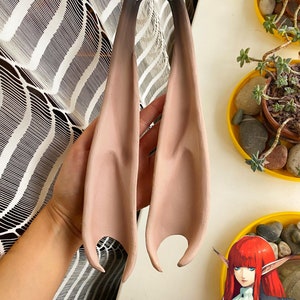 May include: A pair of prosthetic elf ears, flesh-toned with dark brown tips. The ears are held up against a white surface, with a patterned curtain in the background. The ears are designed to be worn and add a fantasy element to a costume.