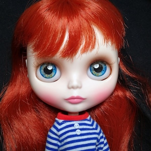 May include: A doll with long, bright red hair, blue eyes with yellow accents, and a striped blue and white shirt. The doll has a light pink blush on its cheeks.