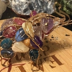 May include: A handcrafted elephant sculpture made of gold-colored wire, filled with polished gemstones in various colors. The gemstones include blue, purple, red, and white stones. The sculpture is displayed on a wooden surface.