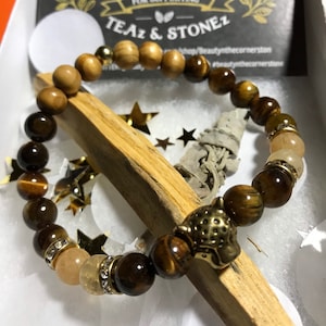 Pode incluir: Um bracelete de contas de olho de tigre castanho e dourado com um fecho de pantera em metal dourado. O bracelete está sobre uma superfície de madeira com um pau de sálvia branca e estrelas douradas.
