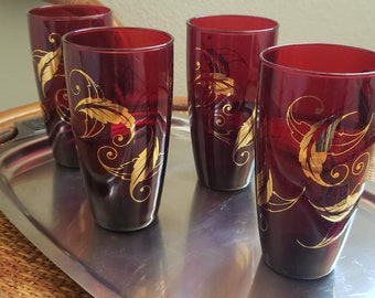 May include: Four tall, ruby red drinking glasses with gold leaf designs. The glasses are cylindrical and sit on a silver metal tray. The gold leaf pattern features swirling lines and leaf shapes.