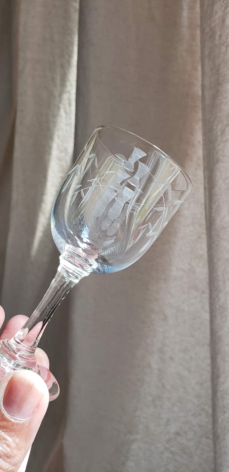 May include: A clear glass stemmed drinking glass with an etched design of bamboo stalks. The glass is held in a hand.